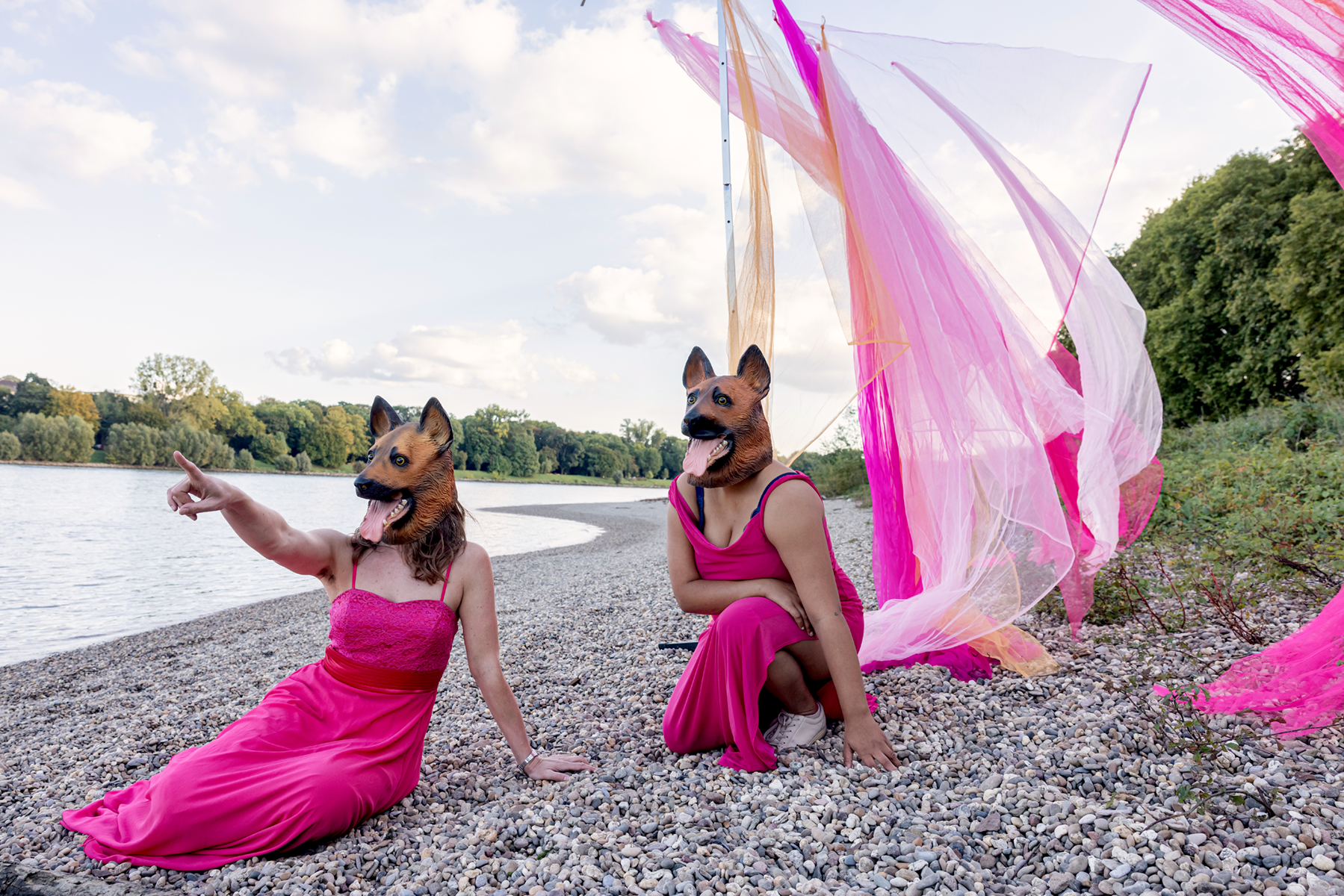 Zwei Personen in pinken Kleidern, die Hundemasken aufhaben und am Ufer eines Flusses sitzen