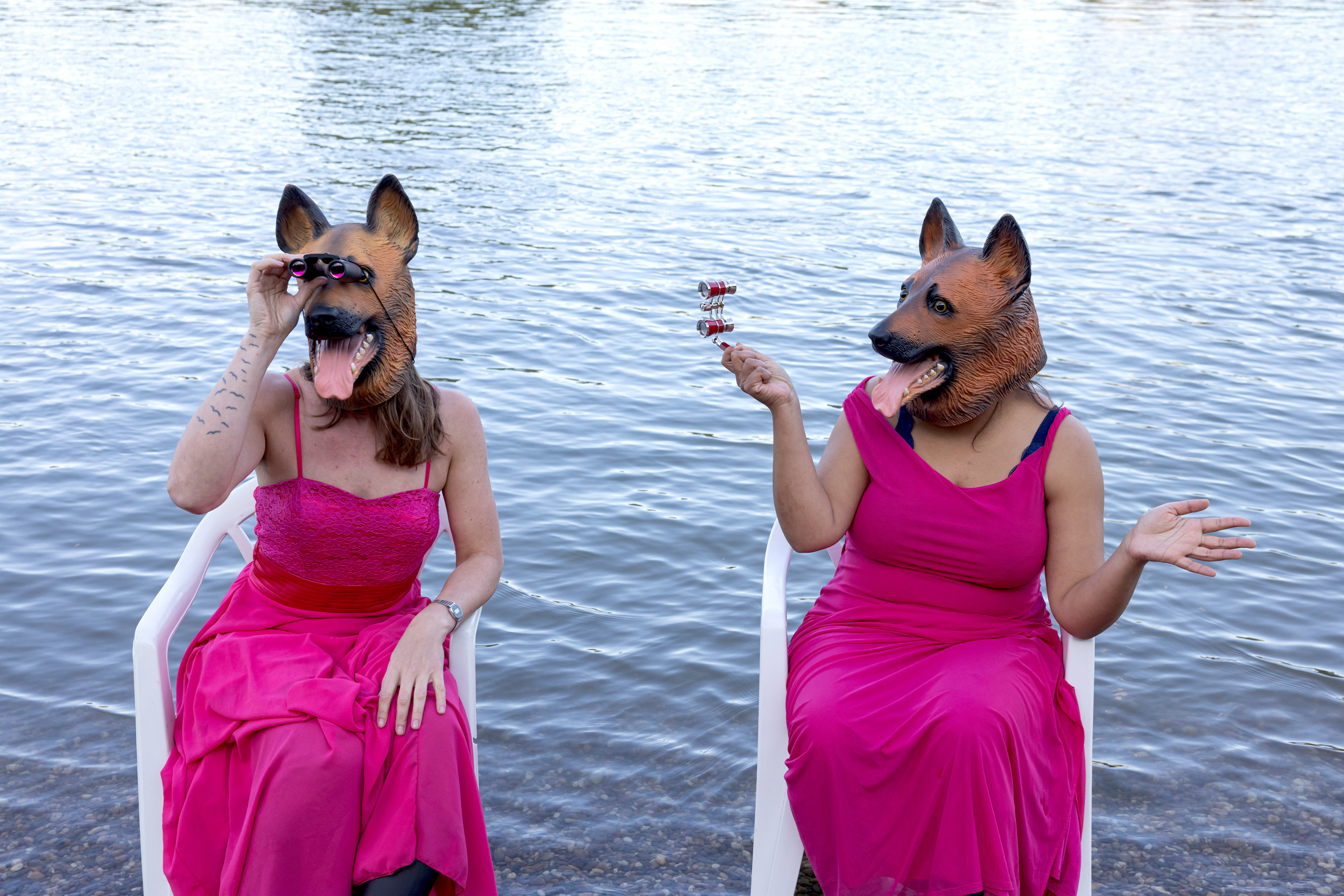 Zwei Personen in pinken Kleidern mit Hundemaske sitzen vor einem Fluss.